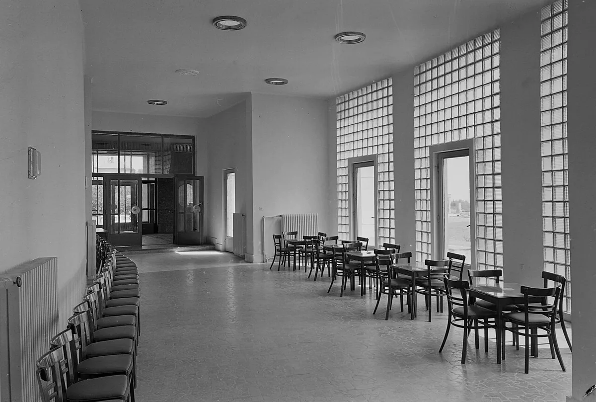 Une salle d'attente vide en noir et blanc : une longue rangée de chaises le long d'un mur de pavés de verre.