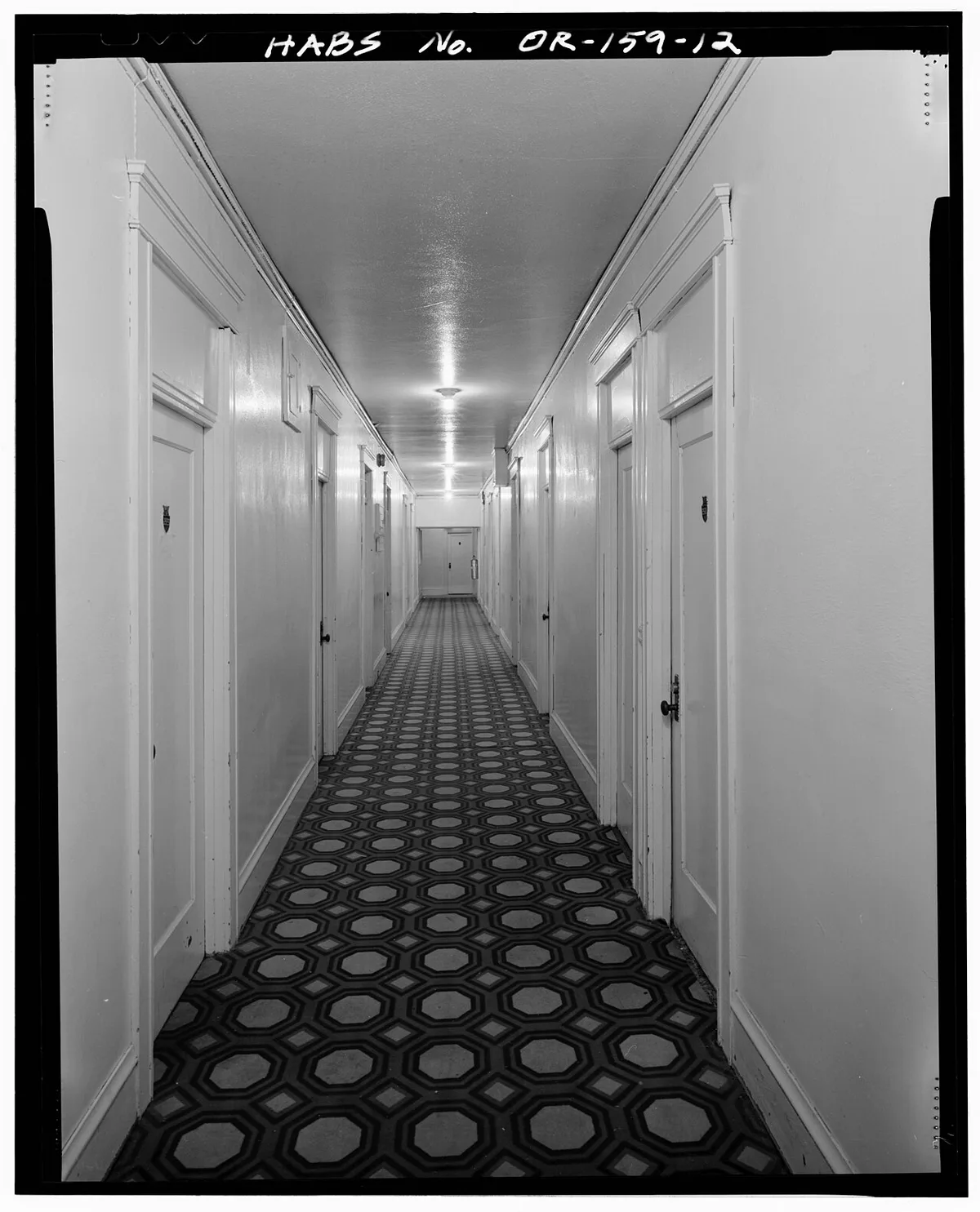 Couloir d'hôtel ancien en enfilade, avec des portes numérotées de part et d'autre et un plancher de bois.