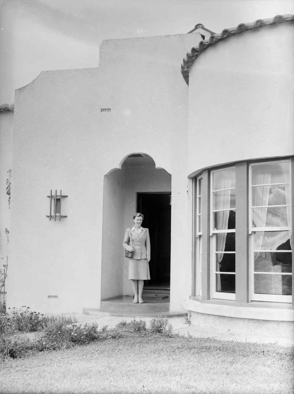 Photographie ancienne en noir et blanc : une femme se tient dans l'encadrement d'une porte cintrée, à côté d'une fenêtre en bow-window.