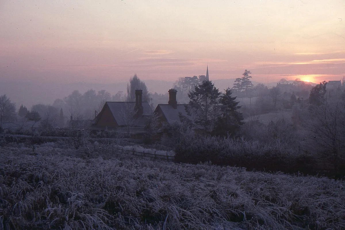 Un village couvert de givre au coucher du soleil, un clocher se détachant sur un ciel rose et brumeux.